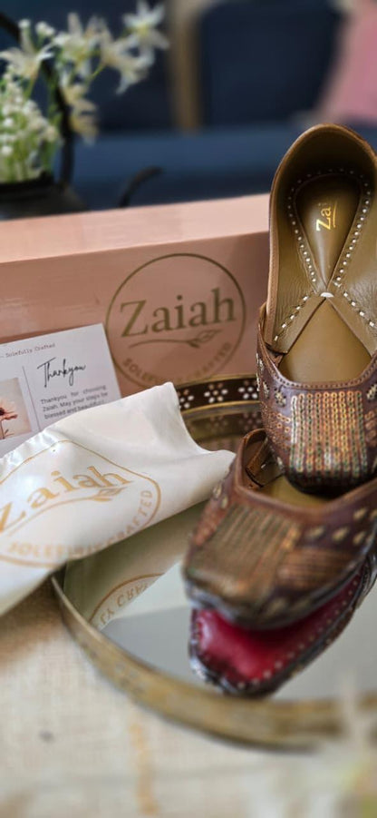 Pair of bronzebloom jutti with 'Zaiah' branding on a reflective surface.